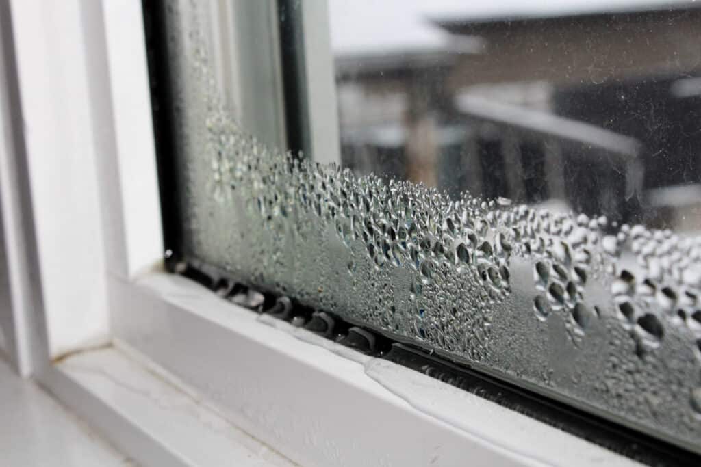 Close-up of a window with a white frame, showing condensation on the inside pane. The water droplets are gathered at the bottom of the glass, indicating humidity or temperature differences between the inside and outside. The slightly blurred background raises a question: does AC lower humidity effectively?