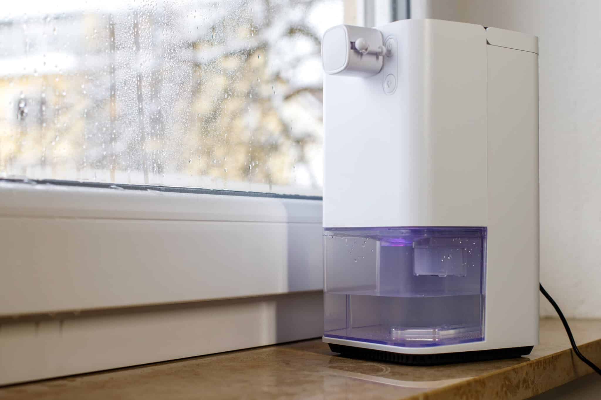 A compact white humidifier with a clear water tank is placed on a windowsill indoors. The window in the background shows droplets and a blurred view of the outdoors, indicating a rainy day. Considering how does a dehumidifier work, this scene captures its opposite—a device adding moisture to the air.