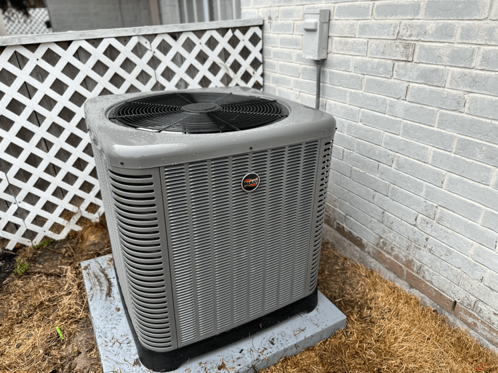 A large gray outdoor air conditioning unit, one of the best AC systems on the market, sits on a concrete slab beside a light gray brick wall. The unit has a fan on the top and is surrounded by a white lattice fence on one side. The ground around the unit is covered in dry grass.