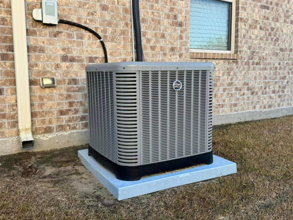 An outdoor air conditioning unit, one of the best AC systems available, is installed on a concrete pad next to a brick house. The unit has a metallic finish with a grid design and is connected to the house via black tubing. Grass surrounds the pad, and a window is visible in the background.