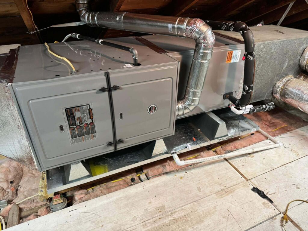 A residential attic with exposed wooden beams showcases an installed HVAC system, boasting one of the best AC systems available. The setup includes a furnace and insulated ductwork. Electrical wires are visible, and some loose insulation is scattered around on the floor.