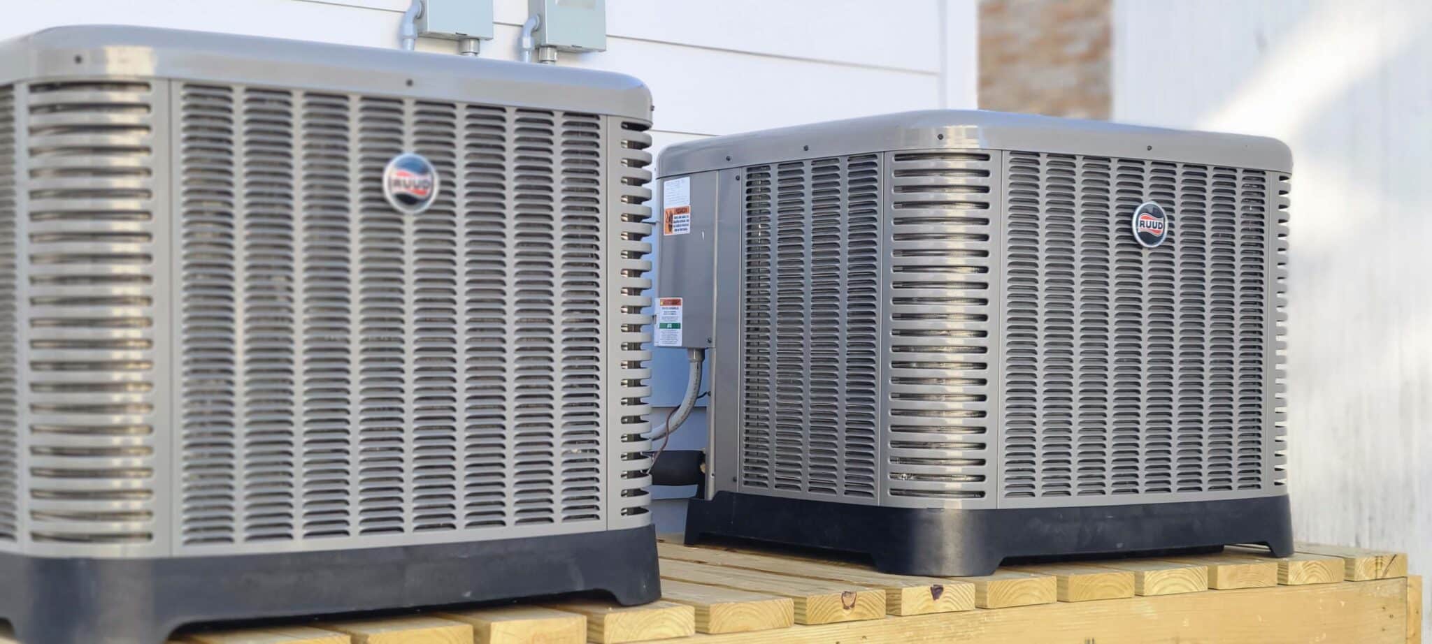 Two grey outdoor air conditioning units, known as the best AC systems, are installed on a wooden platform. Placed side by side near the exterior of a building, electrical conduits run from these top-rated units to the building.