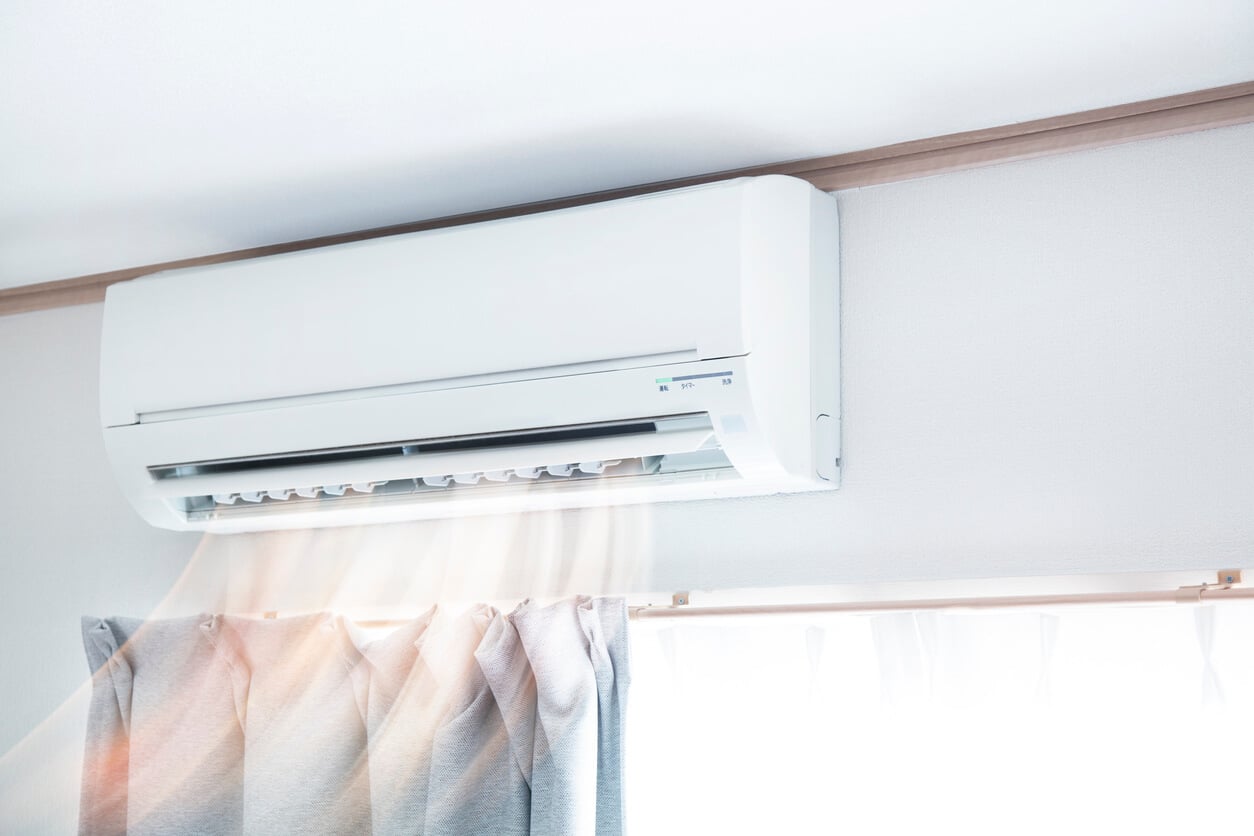 A wall-mounted air conditioner, showcasing how mini splits work, releases cool air above a window with light-colored curtains. Sunlight streams in through the window, creating a bright and airy environment. The airflow is visible as soft, wavy lines below the unit.