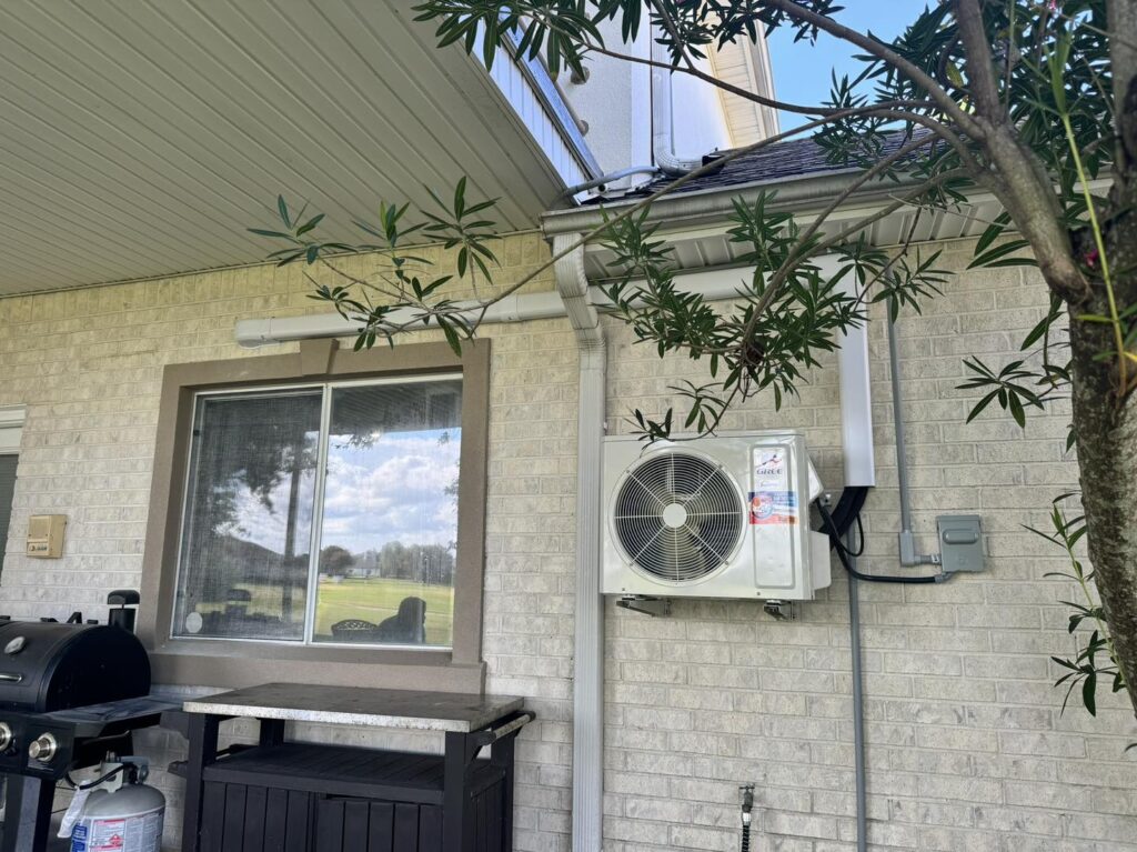 Outdoor view of a residential home with a mini-split air conditioning unit mounted on a brick wall beneath a roof overhang. A window is to the left of the unit, and a barbecue grill stands below the window. For those wondering, "how do mini splits work," efficiency meets aesthetics here. A tree partially obscures the right side of the image.