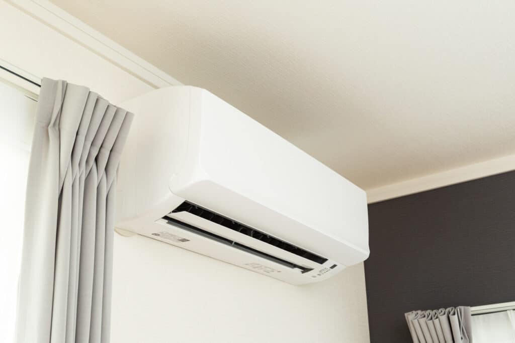 A white, wall-mounted air conditioning unit is installed near the ceiling of a room with neutral-colored curtains. The AC unit appears modern, with a clean and sleek design, blending in with the light-colored walls. Wondering how do mini splits work? This model efficiently cools the space while remaining unobtrusive.