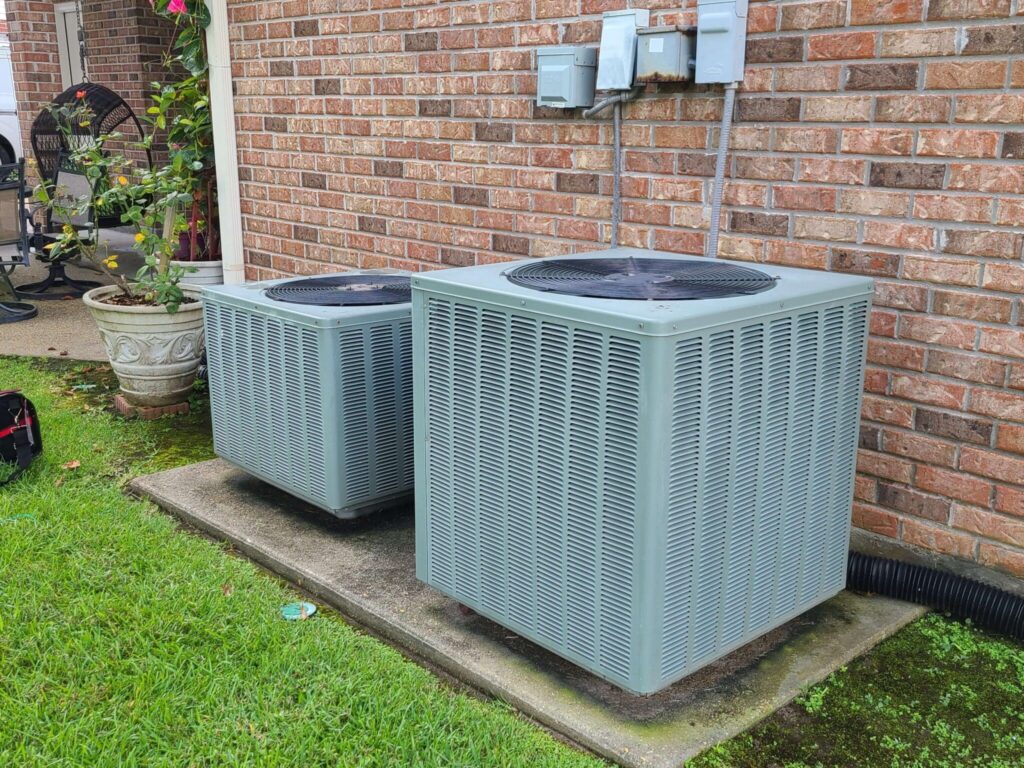 Two outdoor air conditioning units, potential signs of a bad AC compressor, sit on a concrete pad next to a red brick wall. Green grass and plants with pink flowers surround the area, while a small patio chair is partially visible in the background.