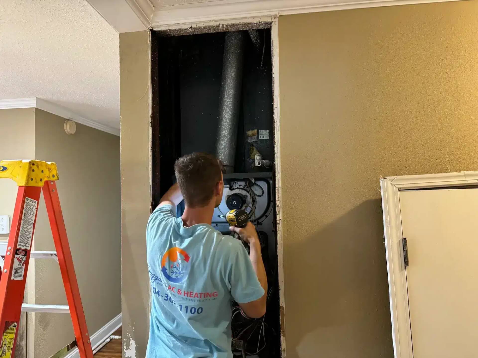 Furnace Service New Orleans, A technician is working on an HVAC unit inside a wall of a residential space. He's using a drill while standing next to a red ladder. The wall is partially open, exposing the unit, and a door leans against the nearby wall.