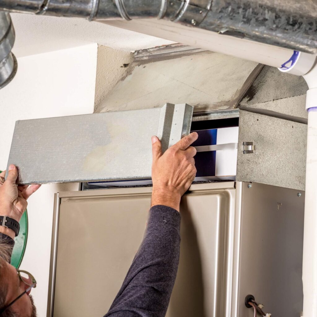 A person is engaged in regular furnace maintenance by changing the air filter of a home HVAC system. The metallic unit features exposed ductwork overhead. Hands grasp a rectangular metal panel, removing it to access the filter inside—a crucial task done as often as recommended to ensure efficiency.