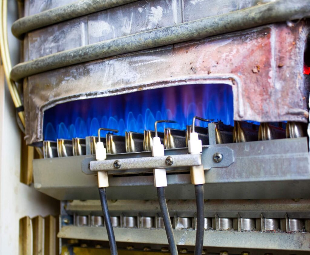 Close-up of a lit gas burner, showcasing a row of blue flames within a metal housing. The burner is part of an appliance with visible components, like pipes and wires, reminding one how often furnace maintenance ensures safe and efficient operation for heating or cooking equipment.