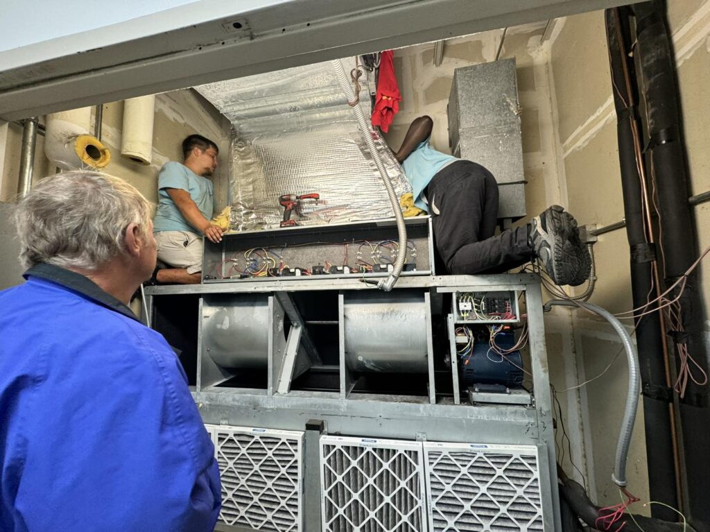 Three people are working on HVAC equipment in a building. Two are kneeling on the unit making adjustments, while the third observes. With wires and tools scattered around and unfinished walls in the background, it's clear who to call for furnace repair if problems arise.