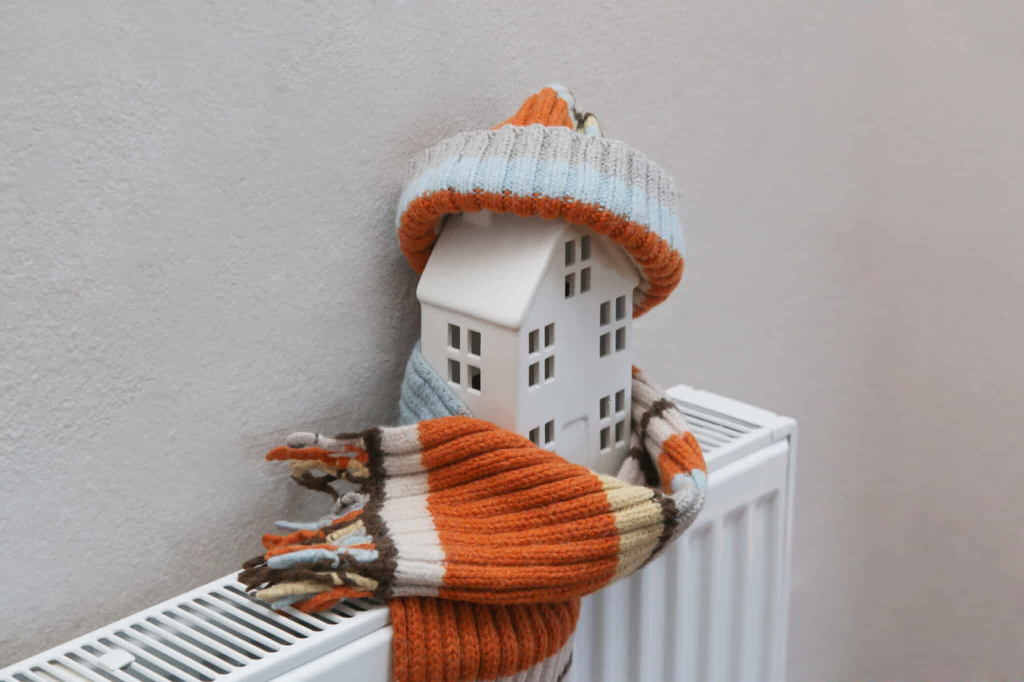 A small white model house sits on a radiator, wrapped in a striped orange and beige scarf, topped with a matching beanie. This cozy setup begs the question: who to call for furnace repair when you need that extra warmth during cold weather?