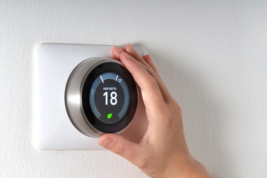A hand adjusts a smart thermostat mounted on a white wall, with the digital display showing a temperature setting of 18°C and a green leaf icon.