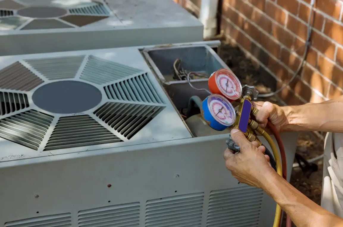A person uses a manifold gauge set to check the pressure on an outdoor air conditioning unit next to a brick wall.