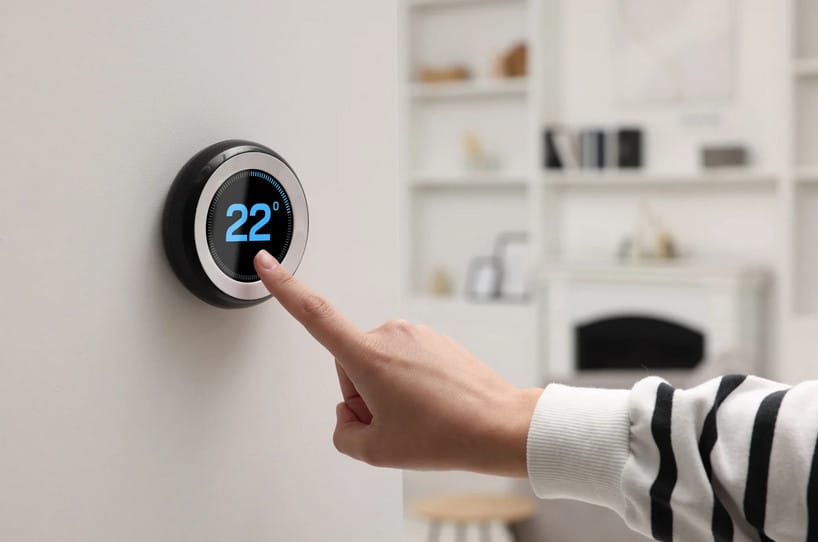 A person wearing a striped sweater adjusts a smart thermostat on a wall, setting the temperature to 22 degrees Celsius. A living room with shelves and a fireplace is blurred in the background.