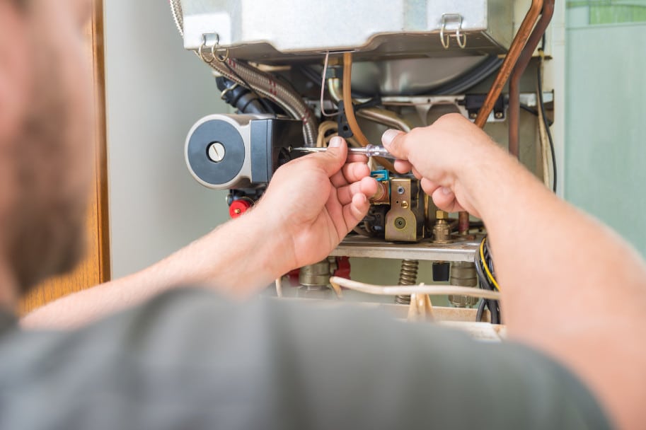 A person uses a screwdriver to repair or maintain the inside of a boiler or heating unit, with various wires and components visible. The image is focused on the hands and the equipment.
