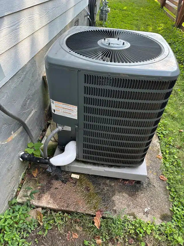 A gray outdoor air conditioning unit sits on a concrete pad next to a house, surrounded by some grass and weeds. Pipes and electrical lines connect the unit to the house's exterior wall.