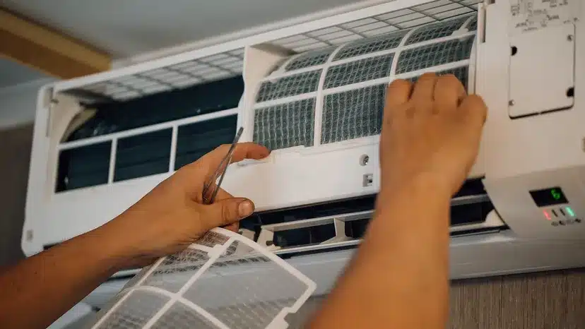 A person is removing and cleaning air filters from a wall-mounted air conditioner unit. The front panel is open, and both hands are handling the mesh filters.