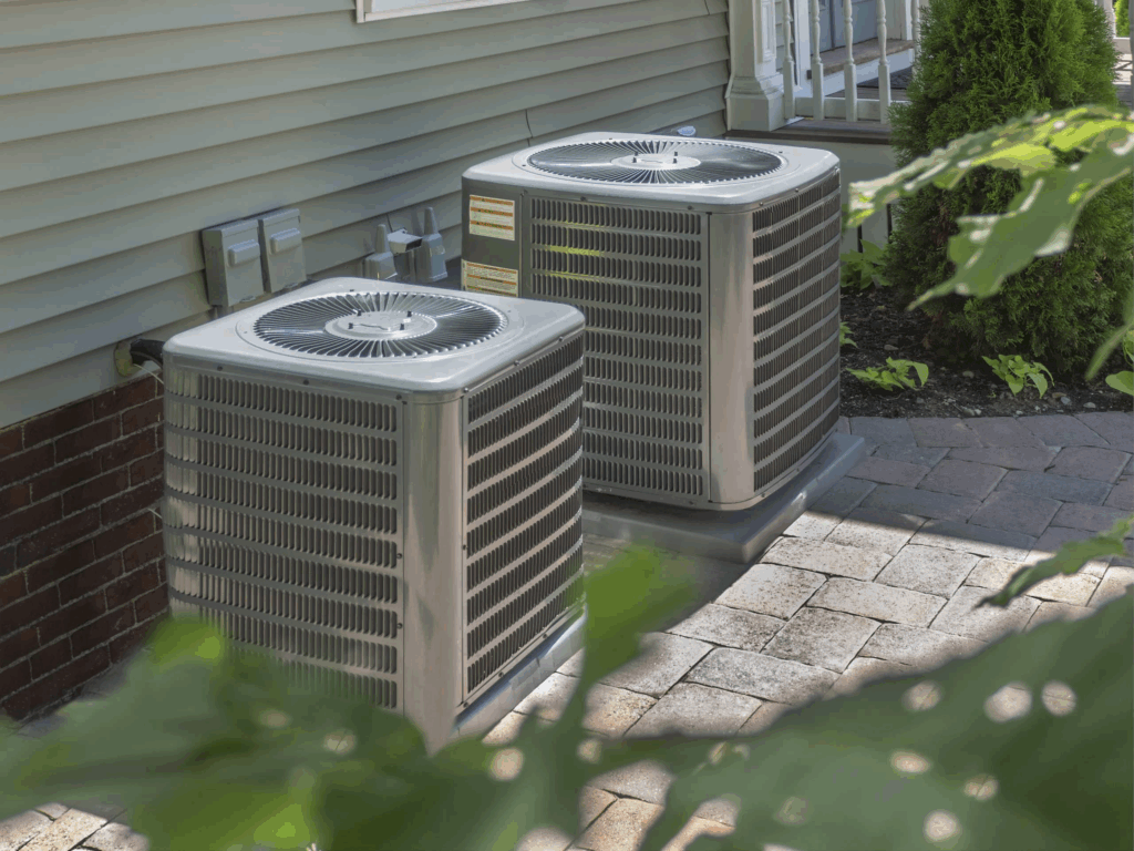 Two outdoor air conditioning units sit on a paved area next to the exterior wall of a house, surrounded by greenery and landscaping.