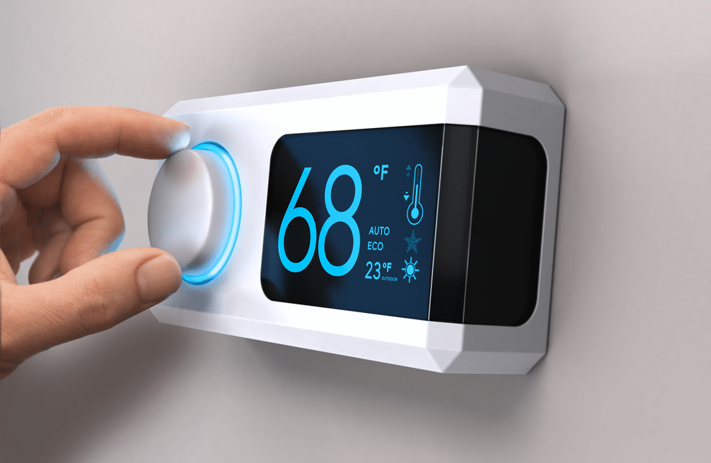 A hand adjusts a modern digital thermostat mounted on a wall. The screen displays the current temperature as 68 degrees Fahrenheit with additional settings and information visible.